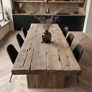 Pode incluir: Uma grande mesa de jantar rústica de madeira com uma aparência natural e desgastada. A mesa é suportada por uma base espessa e sólida e rodeada por seis cadeiras escuras. Um vaso de vidro castanho com ramos secos está sobre a mesa.