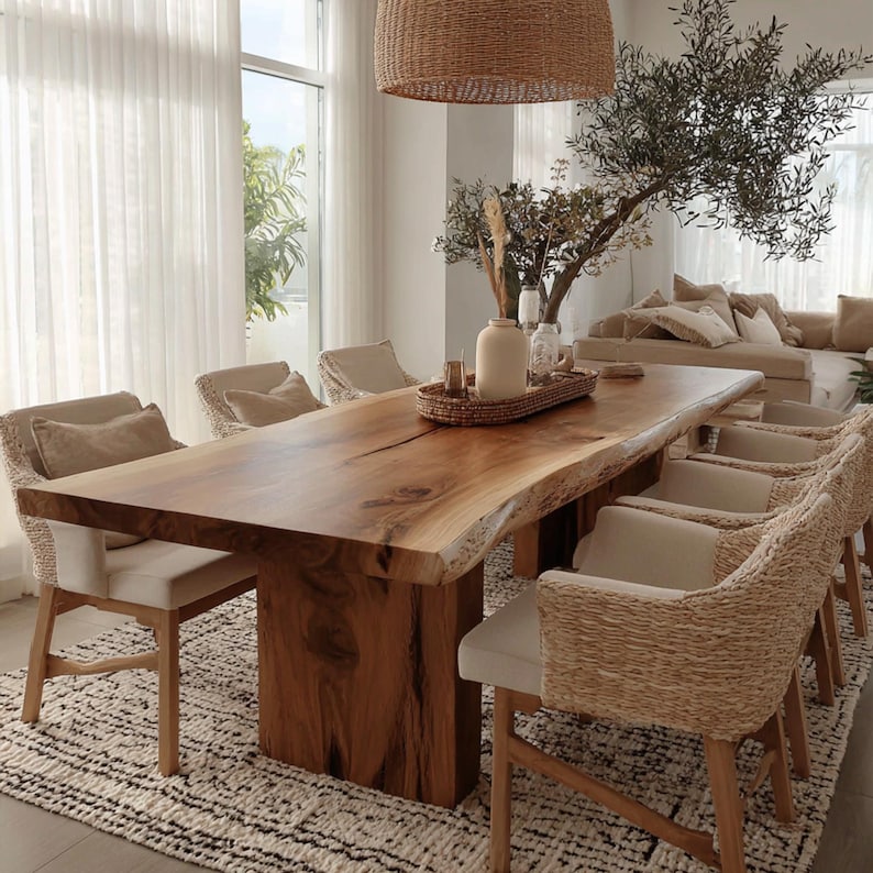 Pode incluir: Uma grande mesa de jantar de madeira natural com borda viva, sustentada por uma base de madeira maci&ccedil;a. Oito cadeiras tecidas com almofadas creme circundam a mesa. O ambiente apresenta uma paleta de cores neutras, uma lumin&aacute;ria tecida e um tapete estampado.