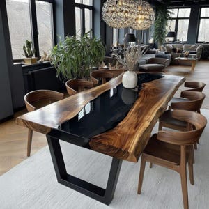 Op de afbeelding: Een grote eettafel met een natuurlijke rand en een zwarte epoxyhars inleg. De tafel heeft een natuurlijke houtnerf afwerking en staat op een zwart metalen onderstel. Rondom de tafel staan houten stoelen met gebogen rugleuningen. De kamer heeft grote ramen en een moderne kroonluchter.