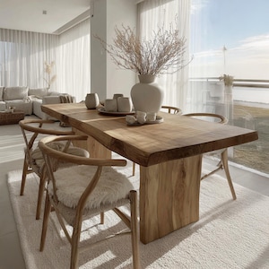 May include: A large, solid wood dining table with a natural edge, supported by a single wooden base. Wooden chairs with curved backs and cushioned seats surround the table. A white vase with dried branches and small ceramic cups and saucers are on the table.