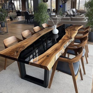 Op de afbeelding: Een grote eettafel met een natuurlijke houten rand en een zwarte epoxyharsrivier in het midden. De tafel wordt ondersteund door een zwart metalen onderstel en is omringd door houten stoelen met zwarte leren zittingen. De tafel is een blikvanger in een moderne eetkamer.