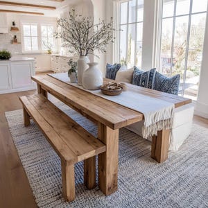 Pode incluir: Uma mesa de jantar rústica de madeira com um banco a condizer, sobre um tapete cinzento texturizado. A mesa é adornada com um caminho de mesa branco, vasos e almofadas decorativas. A sala tem janelas grandes e um ambiente arejado.