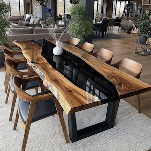 Op de afbeelding: Een grote, rechthoekige eettafel met een houten blad met levende rand en een zwart epoxyhars midden. De tafel wordt ondersteund door een zwarte metalen basis en is omgeven door houten stoelen met zwarte zittingen. De tafel is ongeveer 254 cm lang.