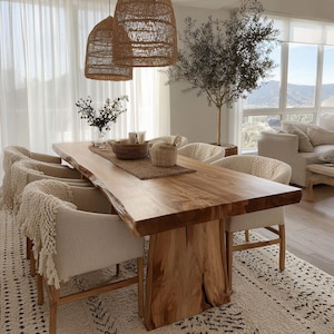 Pode incluir: Uma grande mesa de jantar de madeira natural com borda viva, sustentada por uma base de madeira grossa. Seis cadeiras estofadas com mantas com franjas circundam a mesa. Lumin&aacute;rias pendentes tecidas pendem acima e um tapete estampado est&aacute; por baixo.