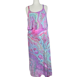 Pode incluir: Um vestido comprido sem mangas com um padrão tie-dye vibrante em tons de magenta, roxo e turquesa. A peça tem um decote redondo e um design de corpete em camadas que cai numa saia longa e fluida com um estampado abstrato consistente.