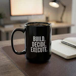 May include: A black coffee mug with the words "BUILD. DECIDE. EXECUTE." in white. The mug is on a brown leather desk mat, next to a notebook and pen. The background includes a desk and a lamp.