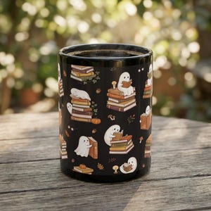 May include: A black ceramic mug featuring a repeating pattern of white cartoon ghosts interacting with stacks of books, small pumpkins, acorns, and lit candles. The ghosts are depicted reading, sleeping on, or hugging the colorful book stacks.