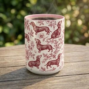 May include: A cylindrical ceramic mug with a light pink interior rim. The exterior features a repeating pattern of dark red dachshund dogs and floral illustrations on a cream-colored background. The mug is filled with a dark liquid.