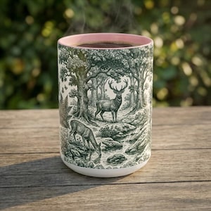 May include: A cylindrical ceramic mug featuring a detailed dark green forest scene with a stag and a deer drinking from a stream. The mug has a soft pink interior rim and a white base, showcasing a woodland illustration with trees and foliage.