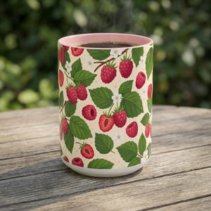 Peut inclure: Un mug cylindrique en céramique avec un fond crème clair orné d'un motif répétitif de framboises rouges, de feuilles vertes et de petites fleurs blanches. Le mug présente un bord intérieur rose pâle et contient une boisson liquide brun foncé.