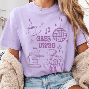 May include: A lilac-colored t-shirt with a graphic that says "Cafe Disco" in a retro font. The design includes a coffee cup, musical notes, a disco ball, and a coffee machine. The model is wearing blue denim shorts and a cream-colored cardigan.