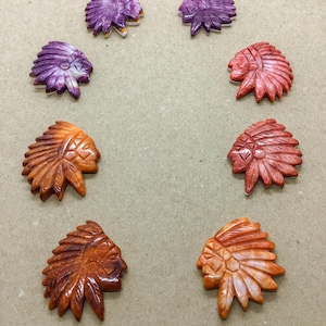 May include: Eight carved cabochons shaped like profiles of individuals wearing feathered headdresses. The pieces are arranged in a circular pattern and feature a variety of colors including shades of purple, red, orange, and brown with a glossy finish.