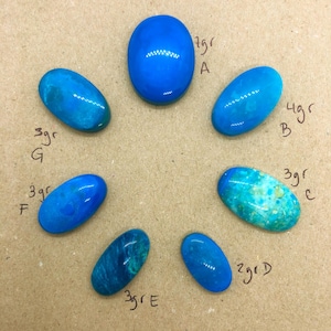 May include: An assortment of seven polished, oval-shaped gemstones in shades of blue and turquoise. The gemstones are arranged in a circular pattern on a neutral-toned surface. Each stone is labeled with a weight in grams.