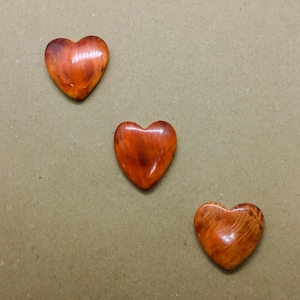 May include: Three heart-shaped objects with a smooth, polished finish. Each piece displays a unique pattern of swirling orange, reddish-brown, and amber tones, resembling the natural grain and texture of polished wood or stone with a glossy sheen.