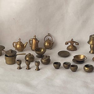 May include: A collection of miniature brass kitchen and dining items. The set includes pots, teapots, bowls, cups, a telephone, a lamp, and plates. The items are arranged on a white surface, showcasing their intricate details and vintage aesthetic.