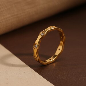 Peut inclure: Une bague dorée au design ondulé. La bague présente de petits ornements scintillants espacés uniformément autour de l'anneau. La bague est posée sur un fond neutre, soulignant son design élégant.