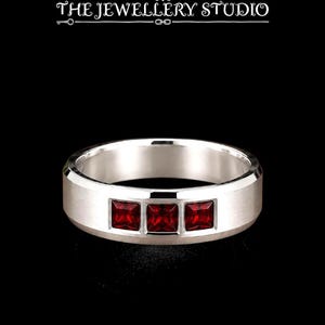 May include: A silver-toned band ring featuring three square-cut deep red gemstones set horizontally in the centre. Above the ring, the text THE JEWELLERY STUDIO is displayed in a white serif font with a small diamond icon above the word THE.