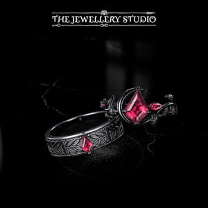 May include: Two rings with dark metal settings and red gemstones. One ring features a large, geometric gemstone and floral accents. The other ring has a band with a leaf pattern and a smaller gemstone. The rings are set against a black background.