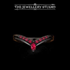 May include: A black V-shaped ring featuring a central marquise-cut red gemstone flanked by three round-cut red gemstones on each side. The text THE JEWELLERY STUDIO is displayed above the ring against a solid black background.