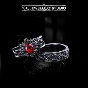 May include: Two rings, one with a large red gemstone and floral accents, the other with a matching floral pattern. The rings are dark gray and set against a black background. The text "THE JEWELLERY STUDIO" is at the top.