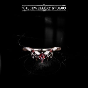 Peut inclure: Une bague en argent avec une pierre centrale en forme de diamant rouge foncé. La bague présente des pierres blanches et des motifs en forme de feuilles de chaque côté de la pierre centrale. Le texte "THE JEWELLERY STUDIO" est en haut.