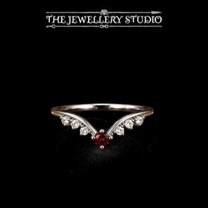 May include: A V-shaped silver-toned ring featuring a central round-cut deep red gemstone flanked by three small clear round-cut stones on each side. The text THE JEWELLERY STUDIO is displayed in white above the ring against a black background.