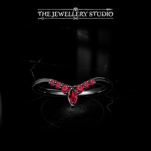 Op de afbeelding: Een zwarte ring met een V-vormig ontwerp, bezet met meerdere kleine, ronde rode edelstenen en een grotere, marquise-geslepen rode edelsteen in het midden. De ring wordt gepresenteerd tegen een zwarte achtergrond. De tekst "THE JEWELLERY STUDIO" staat bovenaan.
