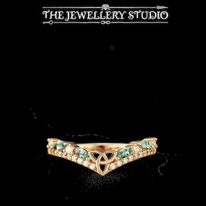 Pode incluir: Um anel dourado com um design em forma de V. O anel apresenta um nó celta no centro, rodeado por pequenas pedras transparentes e pedras preciosas verdes. O texto "THE JEWELLERY STUDIO" está no topo da imagem.