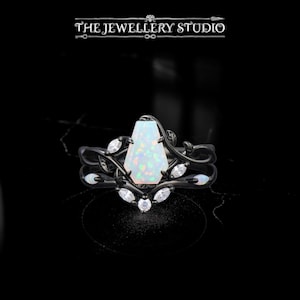 May include: Black ring set with a large, iridescent, rectangular opal center stone. The ring is accented with small, clear gemstones and a delicate, vine-like design. The Jewellery Studio logo is at the top.
