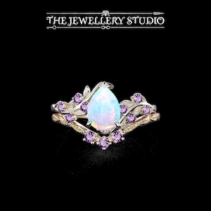 Puede incluir: Un anillo dorado con una piedra central de ópalo en forma de pera y pequeñas gemas moradas. El anillo está rodeado por un diseño de hojas. El texto "THE JEWELLERY STUDIO" está en la parte superior.