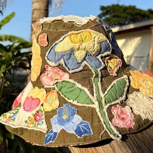 May include: A handmade olive green bucket hat adorned with colorful fabric flower appliqués. The hat features a large central flower with blue and yellow petals, surrounded by various other floral designs in pink, yellow, and white. The brim is trimmed with white lace.