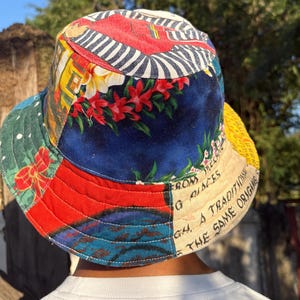 Op de afbeelding: Een kleurrijke patchwork-emmerhoed met verschillende patronen en kleuren, waaronder rood, blauw, groen en geel. De hoed heeft bloemen- en geometrische ontwerpen, met tekst langs de rand. Een uniek modeaccessoire.