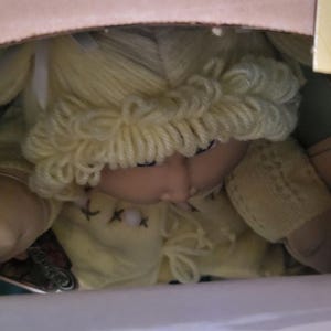 Puede incluir: Una muñeca vintage con cabello de hilo amarillo y un vestido amarillo es parcialmente visible dentro de una caja de cartón. La cara de la muñeca está girada hacia abajo. La caja está abierta, revelando la muñeca y una etiqueta.