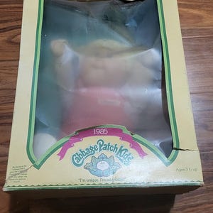 May include: A vintage Cabbage Patch Kids doll in its original packaging from 1985. The box is tan with green trim and features the Cabbage Patch Kids logo and the phrase "I'm unique, I'm adoptable!" The doll is visible through a clear plastic window.