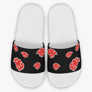 May include: A pair of white slide sandals featuring a wide black strap across the top. The black strap is decorated with a repeating pattern of stylized red clouds with thin white outlines, providing a distinct contrast against the dark background.