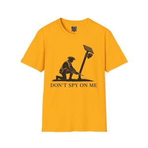 May include: A mustard yellow short-sleeve t-shirt featuring a black graphic of a colonial-era soldier kneeling and using a tool on a post topped with a solar panel and security camera. The text DON'T SPY ON ME is printed below the graphic.