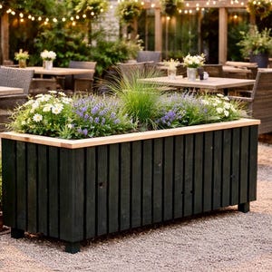 Puede incluir: Una jardinera rectangular de madera verde oscuro, llena de lavanda, margaritas blancas y hierbas. La jardinera tiene un borde de madera clara y está sobre un patio de grava. Mesas y sillas al fondo.