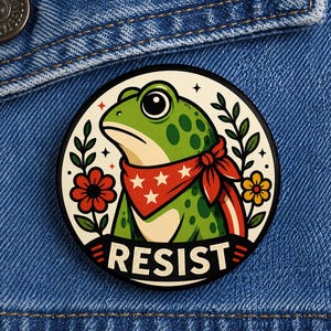 May include: A round button featuring a cartoon frog with a red bandana and the word "RESIST". The frog is green with dark green spots, surrounded by red and yellow flowers and leaves, set against a white background.