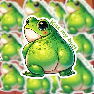 May include: A green frog sticker with the text "Kiss my butt." The frog has large black eyes, a white belly, and a blush on its cheeks. The sticker is surrounded by other frog stickers.