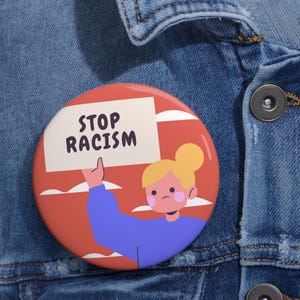 May include: A circular badge with a cartoon design. It shows a person holding a sign with the words "STOP RACISM" on a red and white backdrop. The person is wearing a blue top.