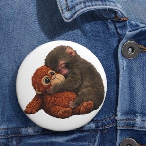 May include: A white button featuring a sleeping baby monkey hugging a stuffed animal. The baby monkey has brown fur and is nestled against a brown stuffed animal with large eyes. The button is set against a blue denim background.