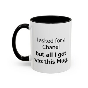 May include: White ceramic mug with a black handle and interior. The mug features the text "I asked for a Chanel but all I got was this Mug" in black, centered on the front.