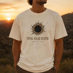 May include: A cream-coloured short-sleeve t-shirt featuring a graphic of a total solar eclipse. The design includes the text "TOTAL SOLAR ECLIPSE", "AUGUST 12 2026", "ICELAND", "SPAIN", and "SAROS 126" with a curved line and small star accents.