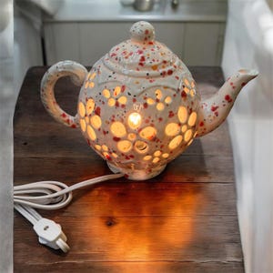 May include: A ceramic teapot-shaped lamp with a cream-colored glaze and red and orange speckles. The body features cut-out flower shapes that emit a warm internal glow. It includes a white electrical cord with a switch and a matching lid.
