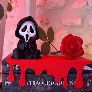 Könnte beinhalten: Eine kleine schwarze Figur mit weißer Geistermaske und rotem Messer sitzt auf einem roten Regal mit tropfendem Design. Daneben liegt eine einzelne rote Rose. Das Regal steht auf zwei Büchern mit den Titeln L'Attaque d'Adeline und L'Ombre d'Adeline.