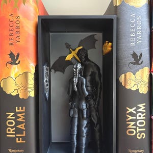 Könnte beinhalten: Eine schwarze Skulptur eines sich umarmenden Paares in einem schwarzen Schaukasten. Ein goldener Drache und ein silbernes Schwert sind an der Rückseite befestigt. Zwei Bücher mit goldener Schrift und Drachenillustrationen flankieren die Präsentation.