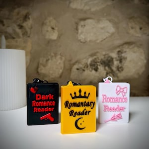 Peut inclure: Trois porte-clés rectangulaires en forme de livre avec des anneaux métalliques. Le noir présente un cœur rouge, un pistolet rouge et le texte "Dark Romance Reader". Le jaune a une couronne noire, un croissant de lune noir et "Romantasy Reader". Le blanc a un nœud rose, une fleur rose et "Romance Reader".