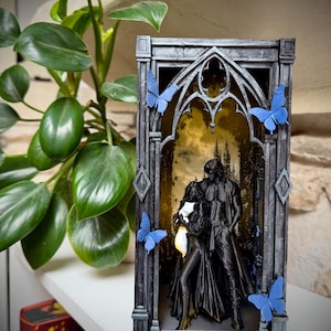 Op de afbeelding: Een rechthoekig, donkergrijs, 3D-geprint gotisch boogframe met een zwart beeldje van twee figuren die samenstaan. Het frame is versierd met vier felblauwe vlinders. De achtergrond in de boog bevat een geelkleurige illustratie.