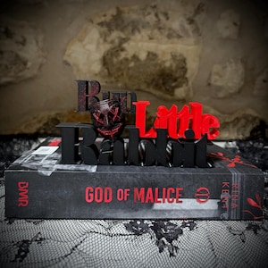 Peut inclure: Un élément décoratif noir et rouge avec l'inscription "Run Little Rabbit" est posé sur un livre intitulé "God of Malice". La pièce décorative comprend un motif de masque et se détache sur un fond texturé.