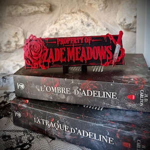 Peut inclure: Un panneau rectangulaire noir avec le texte rouge PROPERTY OF ZADE MEADOWS, orné d'une rose rouge à gauche et d'un petit couteau à droite, posé sur deux livres intitulés L'OMBRE D'ADELINE et LA TRAQUE D'ADELINE de H.D. Carlton.